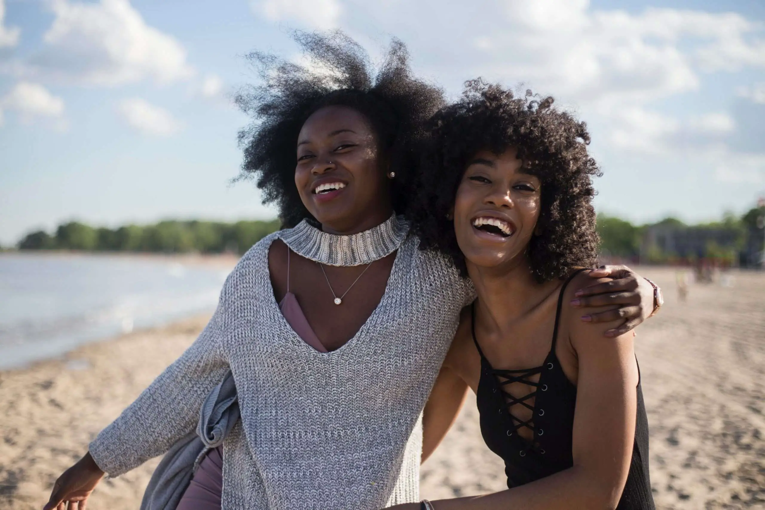 two black girlfriends on the beach on vacation laughing and having a great time. One is wearing grey and the other is black. Both with Afro