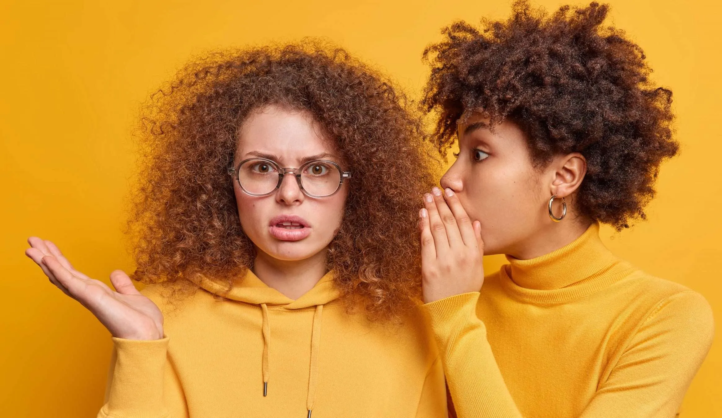 Girl wearing a yellow turtleneck sweater whispering to another girl