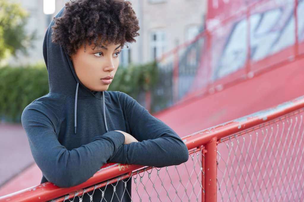 Horizontal shot of thoughtful dark skinned female with Afro haircut, sweatshirt, hood on head, spends free time in playground, has dreamy pensive expression into distance. Active lifestyle concept